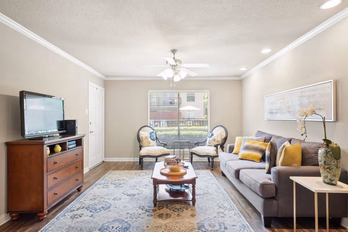 Well-lit living room with a sofa, two accent chairs, a coffee table on a patterned rug, and a TV on a wooden dresser beneath a central ceiling fan.