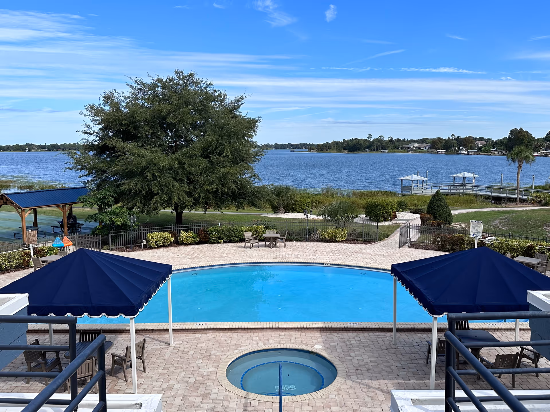 Outdoor pool area with a circular hot tub in the foreground and a larger swimming pool behind it. There are two blue canopy shades with chairs and tables underneath on either side of the pool. Beyond the pool area is a large tree, green shrubs, a walking path, and a lake with a dock and gazebos. The sky is clear with some wispy clouds.