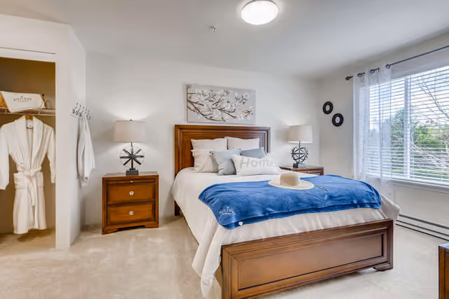 A bright and cozy bedroom featuring a wooden bed with white and blue bedding, two matching wooden nightstands with lamps, a closet with hanging robes, a large window with white blinds, and decorative wall art above the bed.