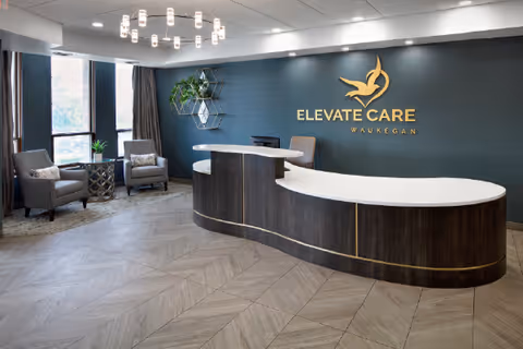Reception area of Elevate Care Waukegan featuring a curved dark wood desk with a white countertop, two gray armchairs with a small round table between them near large windows with curtains, a modern chandelier, and a dark accent wall with the Elevate Care Waukegan logo in gold.