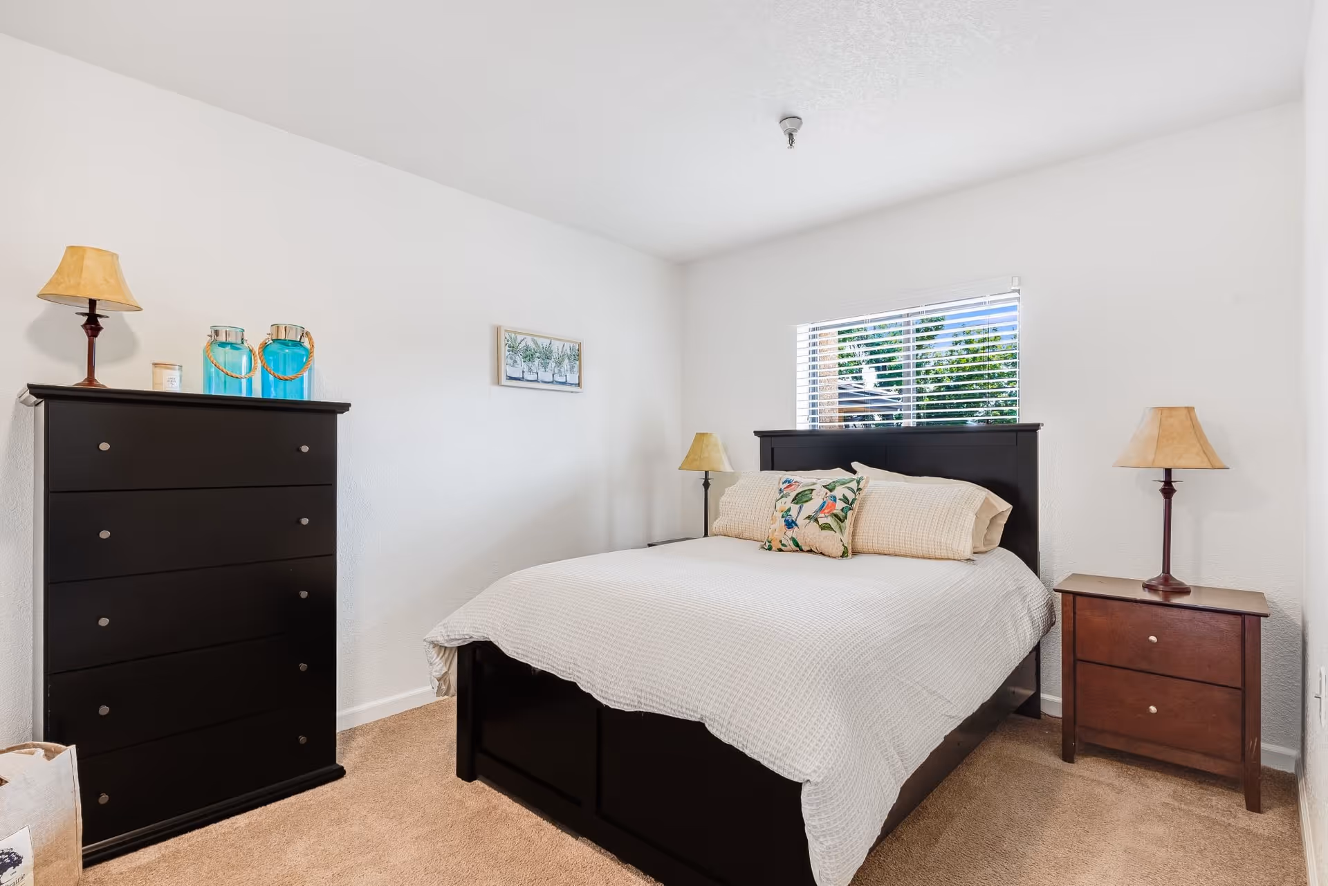 A bedroom with a large bed covered in a white textured blanket and a decorative pillow. The room has beige carpet, white walls, a window with blinds, a dark wooden dresser with two blue glass jars and a lamp on top, and a matching dark wooden nightstand with a lamp.