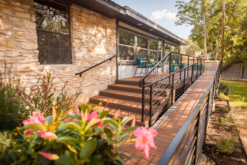 Stone-faced exterior with a wooden ramp and steps leading to a patio with chairs, framed by pink flowers in the foreground.