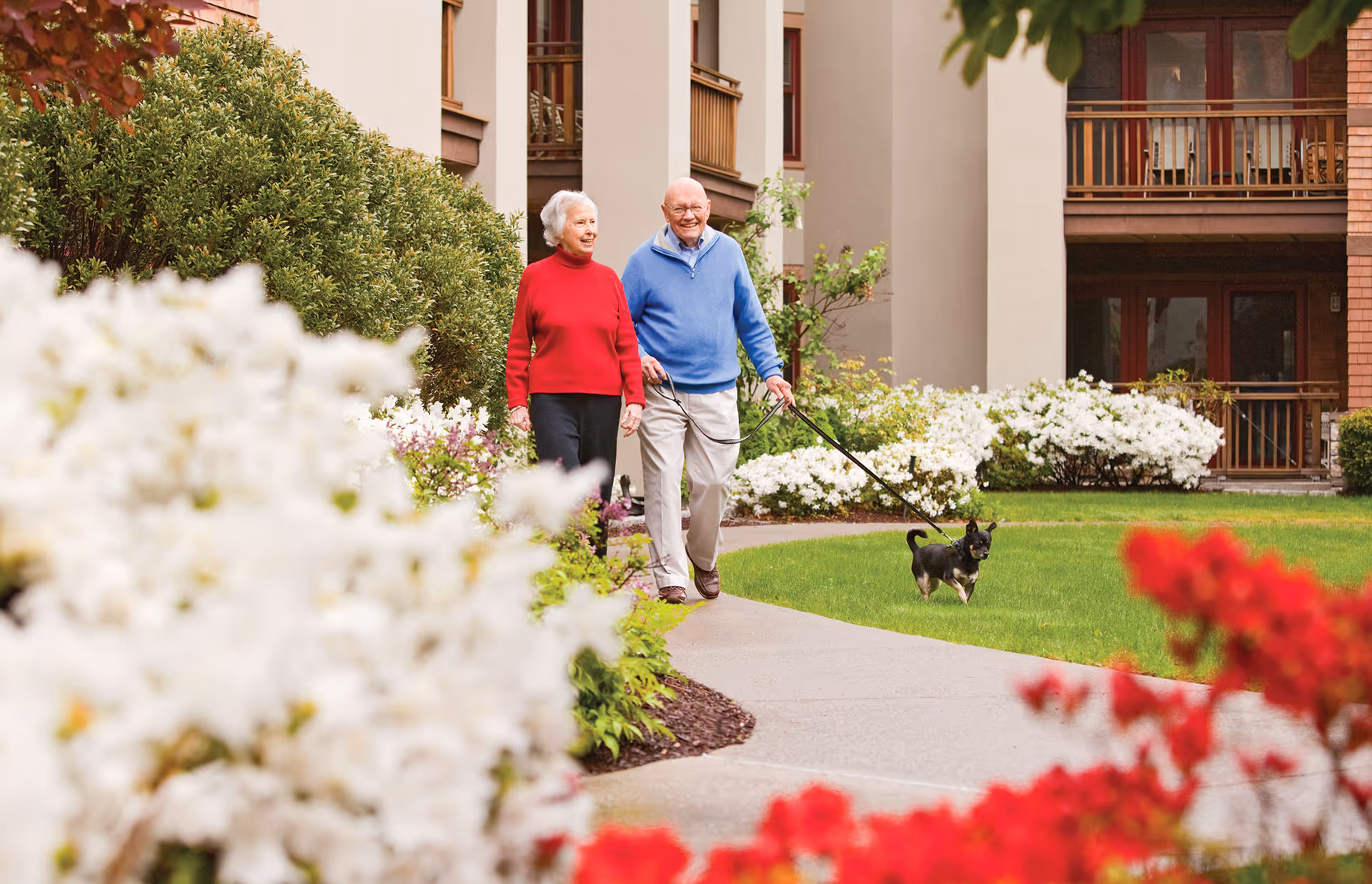 An elderly couple walking a small dog on a leash along a paved pathway surrounded by green grass and blooming flowers in the garden area of a senior living community building.