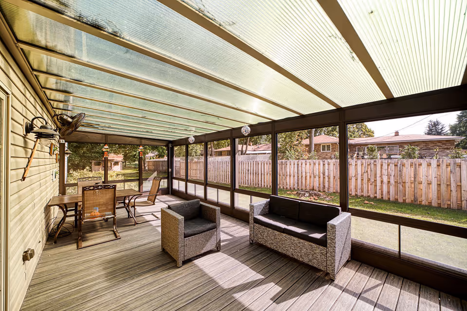 A sunlit enclosed patio with a transparent roof and screened walls. The patio features a wicker armchair and sofa with dark cushions, a wooden table with two chairs, and a wall-mounted fan. Outside the patio is a wooden fence and some greenery.