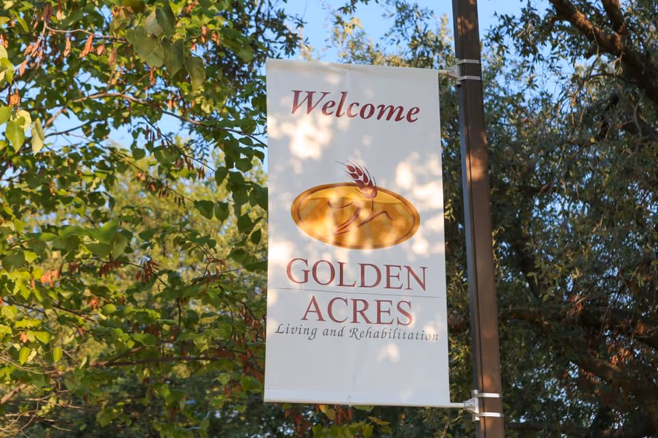 A vertical banner hanging from a pole among green leafy trees. The banner reads 'Welcome GOLDEN ACRES Living and Rehabilitation' with a golden oval logo featuring a stylized wheat design.