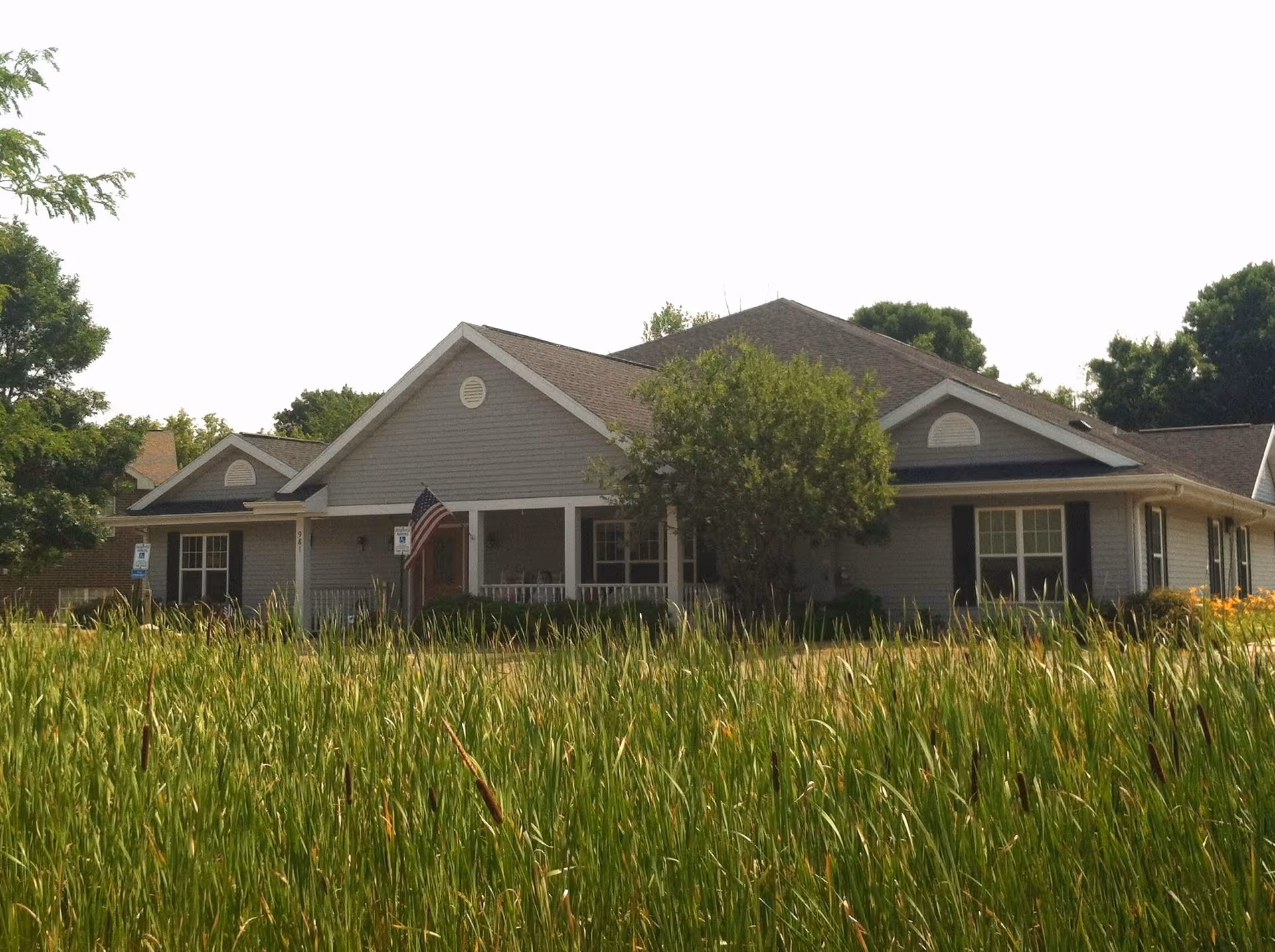 Exterior view of a single-story residential building with gray siding and a gabled roof, partially obscured by tall grass and trees. An American flag is displayed near the entrance, and there are handicap parking signs visible in front of the building.