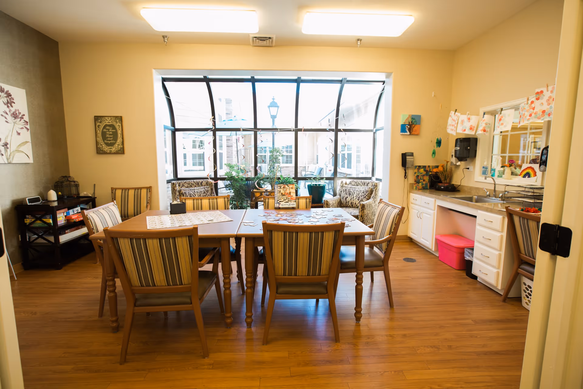 A well-lit room with a wooden table surrounded by six striped cushioned chairs. On the table, there is a partially completed puzzle. The room has a large window with a view of an outdoor area with plants and patio furniture. To the right, there is a small kitchenette area with a sink, cabinets, and a pink storage bin underneath. The walls are decorated with artwork and a small shelf with board games is visible on the left side.