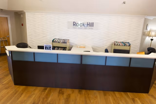 Reception desk area inside Rock Hill Post Acute Care Center with a modern white textured wall behind it, two file organizers filled with folders, a digital clock displaying the time and date, and a lamp on the right side.