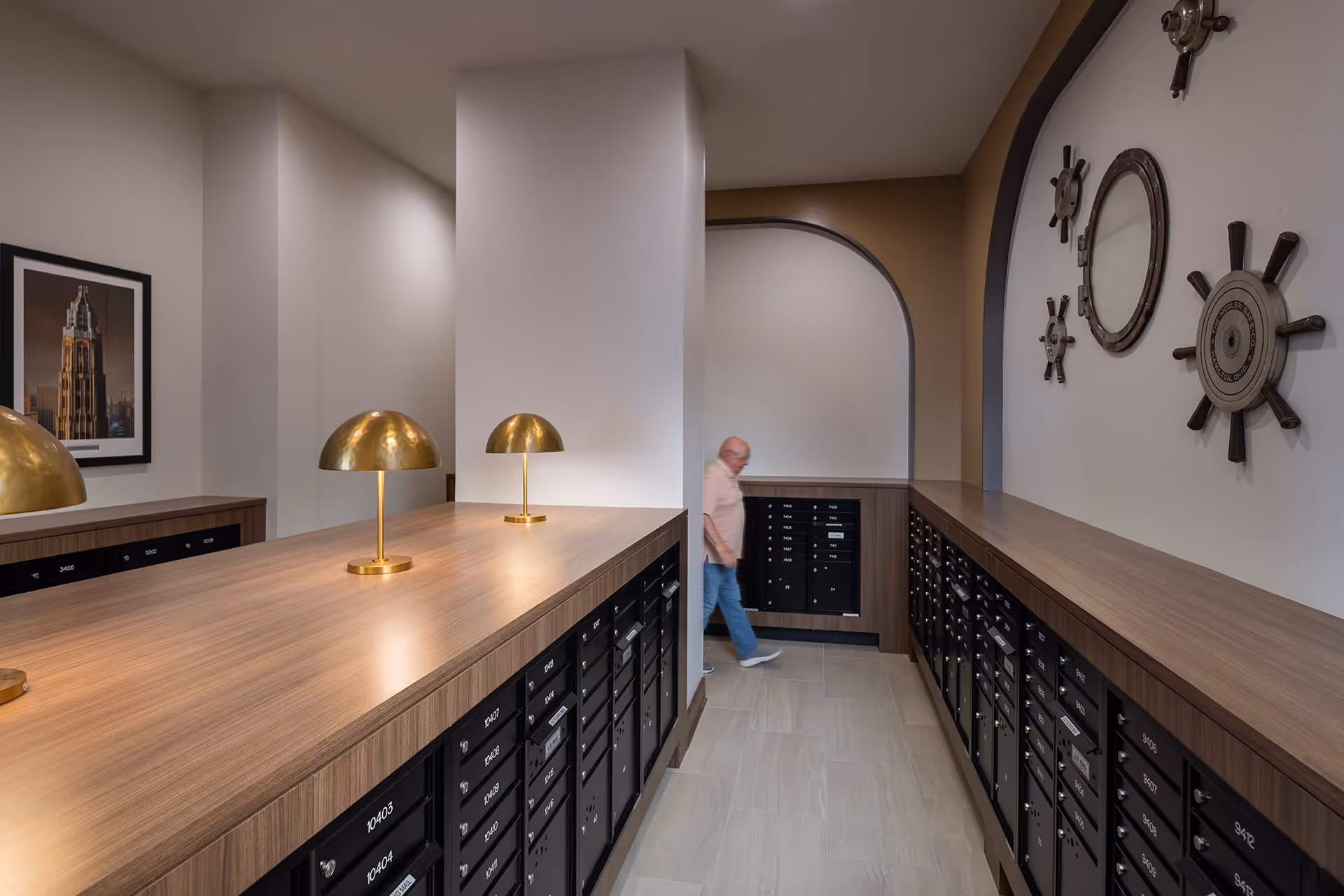 Interior mailroom with rows of wood-topped mailboxes, brass desk lamps, nautical wall decor, and a person walking through an archway.