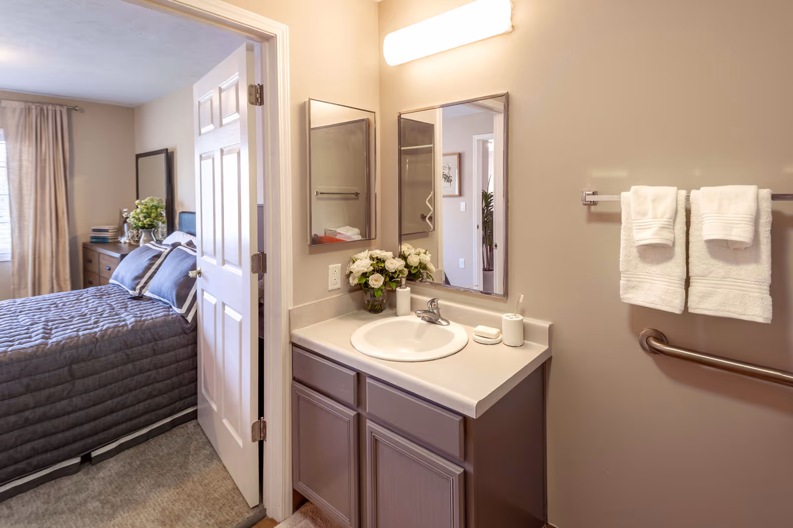 View of a bathroom vanity with a sink, mirror, and light fixture above. Two white towels hang on a towel rack next to the vanity. Through an open door, a bedroom with a neatly made bed, dresser, and window with curtains is visible.