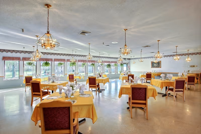 A spacious dining room with multiple tables covered in yellow tablecloths, each set with plates, napkins, and utensils. The room has large windows along one wall with hanging plants and several decorative chandeliers providing lighting. Chairs with red upholstery are placed around the tables.