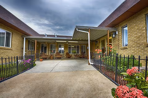 Outdoor courtyard area of a senior living facility with a covered walkway, brick walls, black metal fencing, potted plants, and outdoor seating under a cloudy sky.
