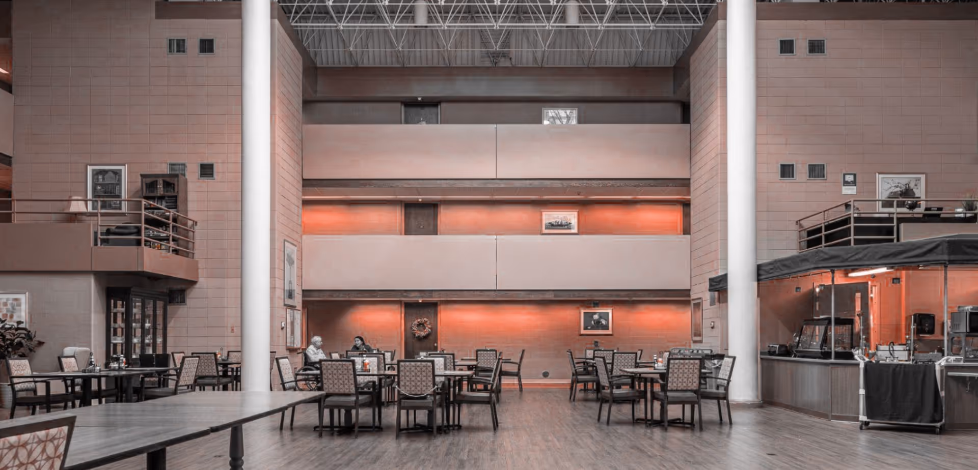 A spacious dining area with multiple tables and chairs arranged neatly. Two people are seated and conversing at one of the tables. The room features high ceilings with exposed beams, large white columns, and a serving station on the right side. The walls are decorated with framed pictures and the lighting has a warm orange hue.