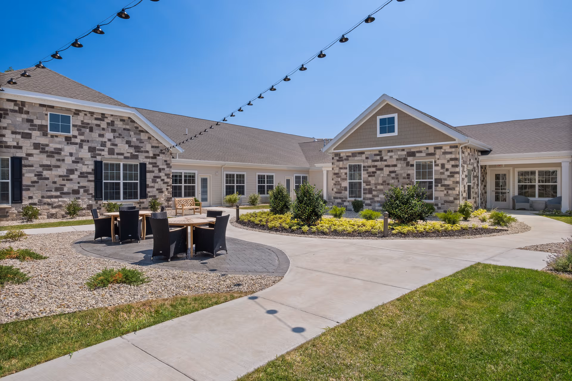 Outdoor courtyard area of Quincy Place Senior Living with stone and siding buildings surrounding a landscaped garden. There are several black wicker chairs and wooden tables on a paved area, string lights overhead, and a clear blue sky.