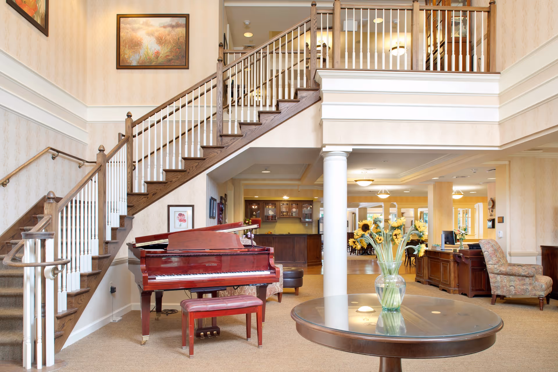 Bright senior living facility lobby with a red grand piano, a round table with a vase of flowers, a staircase, and a reception area.