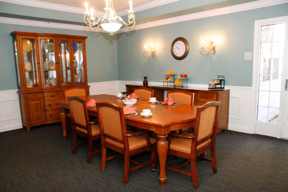 A dining room with a wooden table set for six people with cups, plates, and folded napkins. There are six wooden chairs with cushioned seats around the table. A wooden china cabinet with glass doors displaying dishes is against the left wall. On the right wall, there is a sideboard with a tiered fruit stand and a clock above it. The walls are painted light blue with white wainscoting, and a chandelier hangs from the ceiling. A glass door is visible on the right side of the room.