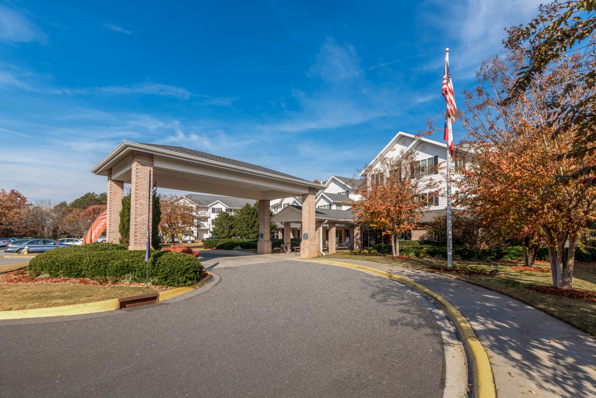 Covered entrance/porte-cochere of a senior living building with a curved driveway, American flag, and autumn trees.
