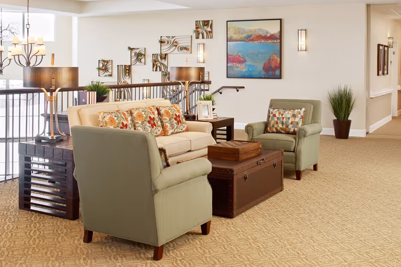 A cozy seating area in a senior living facility featuring a beige sofa with floral cushions, two green armchairs with patterned pillows, a dark wooden coffee table resembling a trunk, and side tables with lamps. The background includes a railing, decorative wall art, a landscape painting, and potted plants on a carpeted floor.