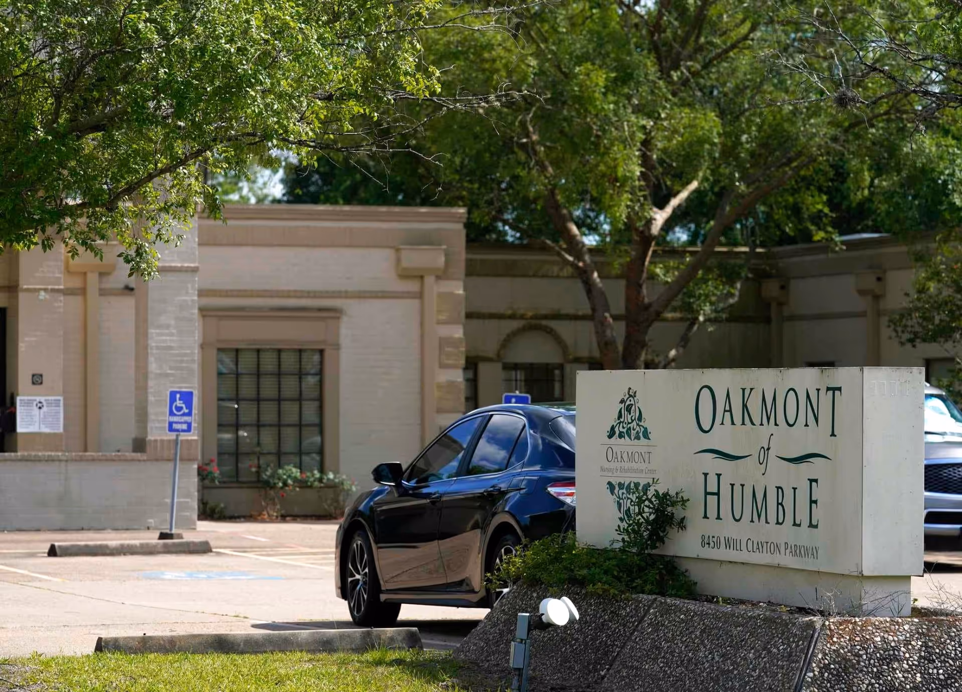 Exterior view of Oakmont Healthcare & Rehabilitation Center of Humble showing the facility's sign with address 8450 Will Clayton Parkway, a black car parked nearby, and trees providing shade.
