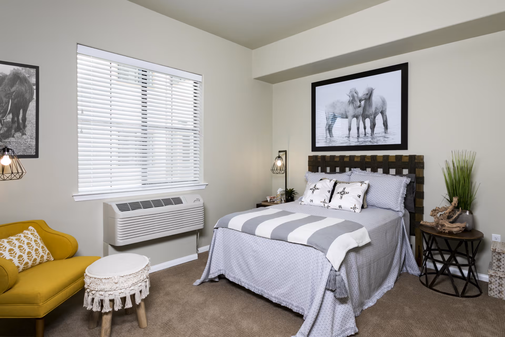 A cozy bedroom with a bed covered in gray and white bedding, two decorative pillows, and a striped throw blanket. Above the bed is a framed black and white photo of two horses. To the right of the bed is a small round table with a plant and decorative driftwood. To the left is a yellow armchair with a patterned pillow and a small round stool with a fringed cover. A window with white blinds and a wall-mounted air conditioning unit is on the left wall. The room has beige walls and carpeted floor.