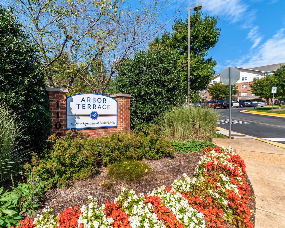 Outdoor view of the entrance sign for Arbor Terrace, a senior living facility, surrounded by greenery and colorful flowers with a building and parked cars in the background under a blue sky.