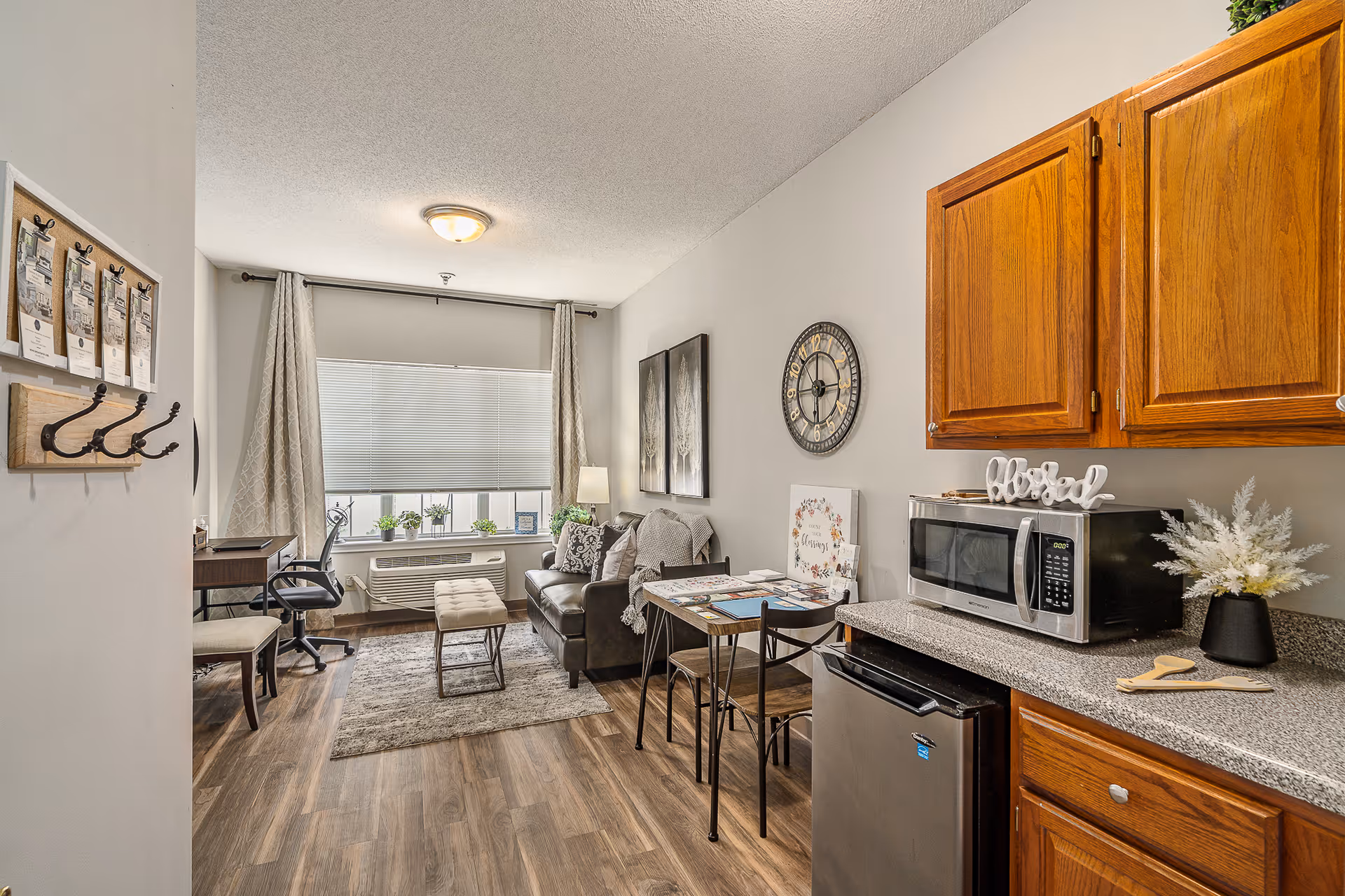 A studio apartment living area with a small kitchenette, sofa, dining table, desk, and a large window.
