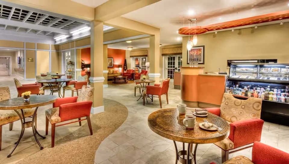 A cozy common area in a senior living facility with round tables and cushioned chairs. The room features warm lighting, a small counter with hanging lights, and a refrigerated display case stocked with beverages and snacks. The space has a mix of tiled and carpeted flooring, with a seating area visible in the background.