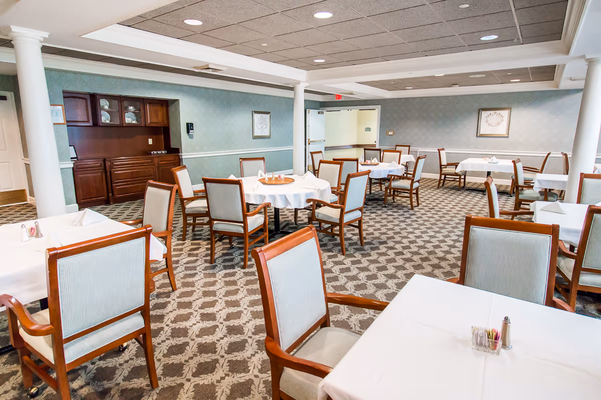 A dining room in a senior living facility with multiple tables covered with white tablecloths and surrounded by wooden chairs with light-colored upholstery. The room has patterned carpet, light blue walls with white trim, and a built-in wooden cabinet with dishes. There are framed pictures on the walls and ceiling lights providing illumination.