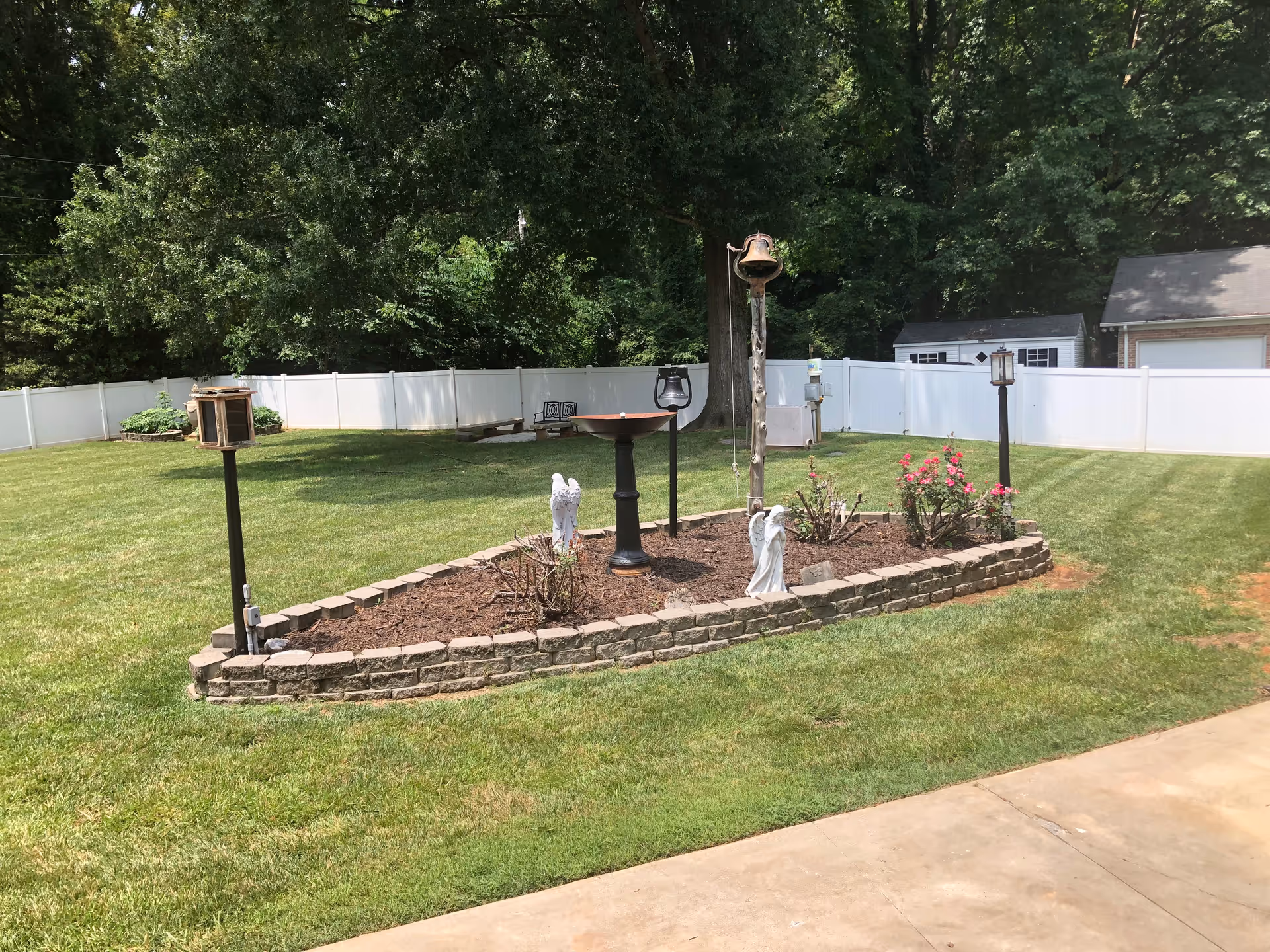 A well-maintained outdoor garden area with a small raised flower bed bordered by bricks. The flower bed contains some plants, two white angel statues, a birdbath, and a tall post with a bell. The garden is surrounded by a white fence, with trees and a small shed visible in the background. The foreground shows a concrete pathway and green grass.