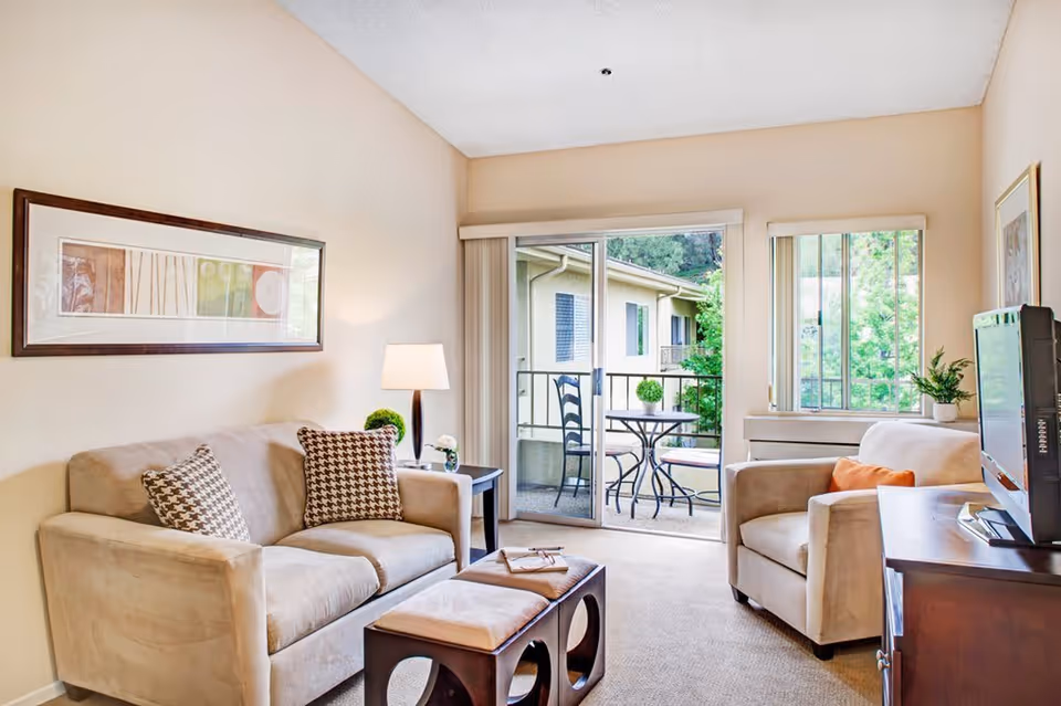 A bright living room with beige walls and carpet, featuring a beige sofa with two patterned cushions, a matching armchair with an orange cushion, a dark wooden coffee table with two cushioned stools underneath, a side table with a lamp and small plants, a flat-screen TV on a wooden stand, and sliding glass doors leading to a small balcony with a round table and two chairs. Outside the balcony, there are trees and neighboring building walls.