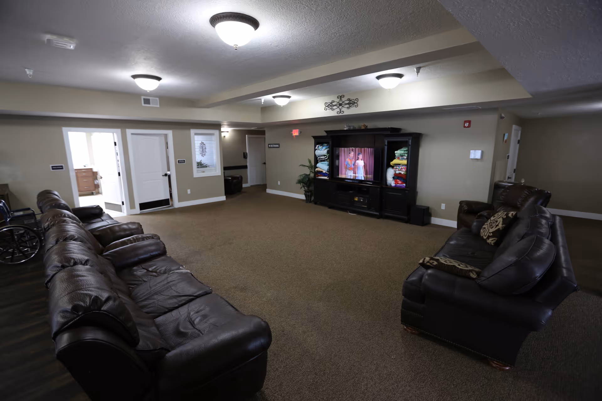 A spacious living room area with brown leather sofas arranged facing a large entertainment center with a television. The TV is on, showing a scene with two people. The room has beige walls, carpeted floor, and ceiling lights. There are several doors and a wheelchair visible on the left side.