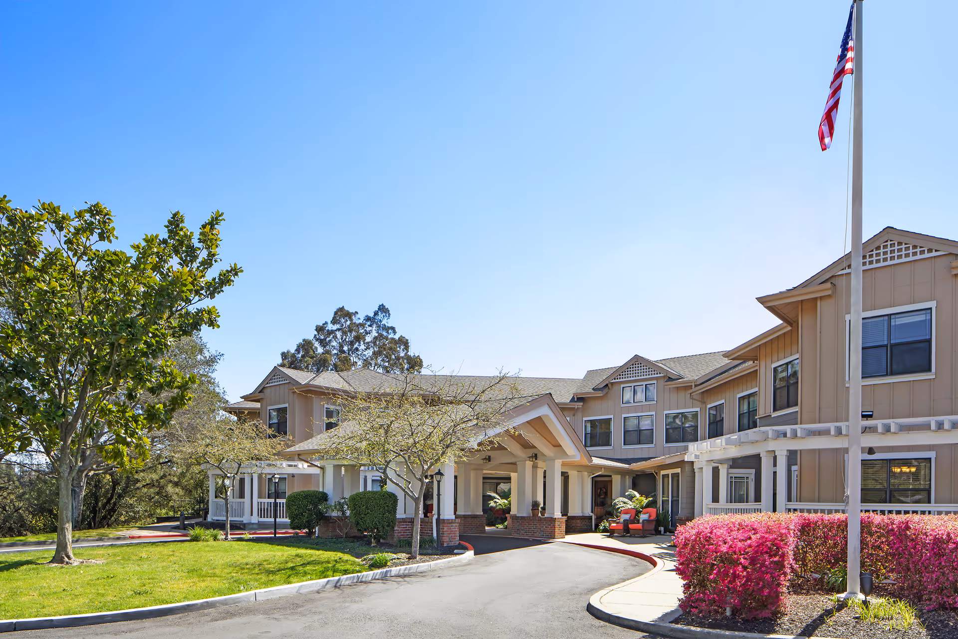 Exterior view of a senior living facility building with beige siding and multiple windows under a clear blue sky. There is a covered entrance with white columns and brick bases, a curved driveway, green grass, trees, and pink flowering bushes. An American flag is flying on a flagpole near the entrance.