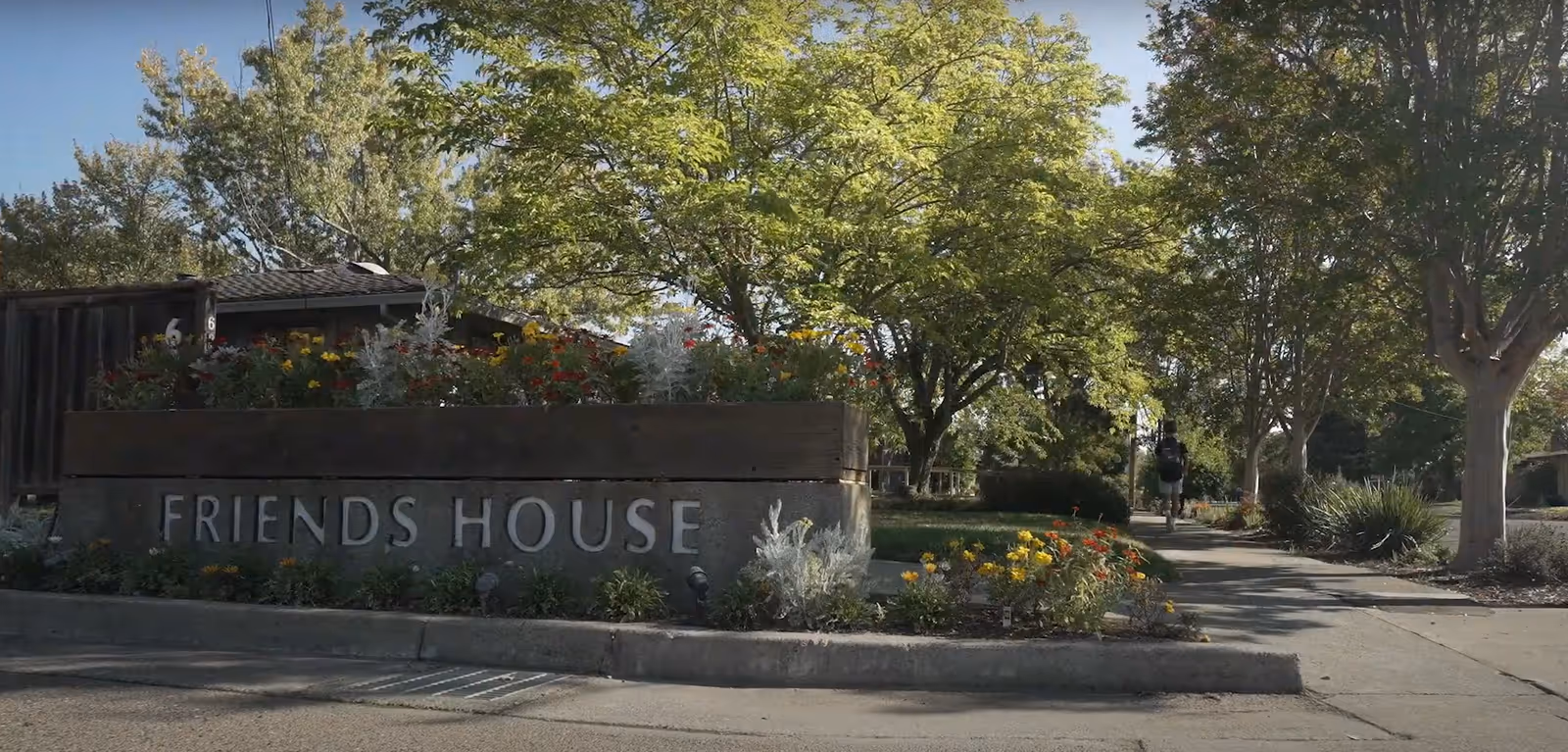 Concrete planter sign reading "FRIENDS HOUSE" at the entrance with flowers, trees, and a walkway.
