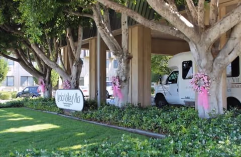 Entrance area of Linda Valley Villa featuring a covered driveway supported by concrete pillars wrapped with pink floral decorations. There is a white vehicle parked under the structure and a sign with the facility's name surrounded by green bushes and trees.