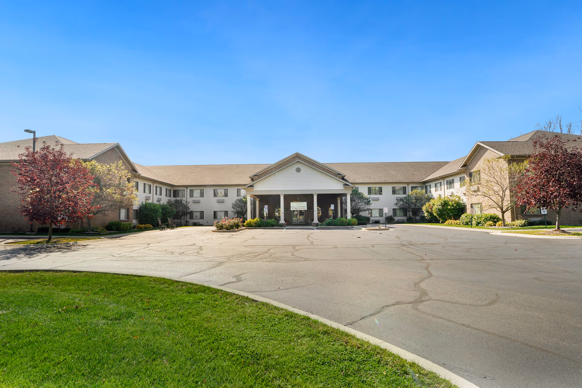 Front exterior view of a two-story senior living facility building with a covered entrance, surrounded by trees and a paved driveway under a clear blue sky.