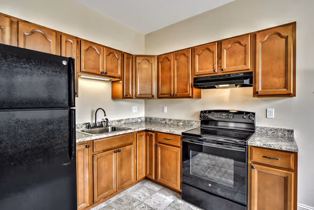 A kitchen with wooden cabinets, a black refrigerator, a black electric stove with an overhead range hood, a stainless steel sink with a black faucet, and granite countertops. The floor is tiled with light-colored tiles.