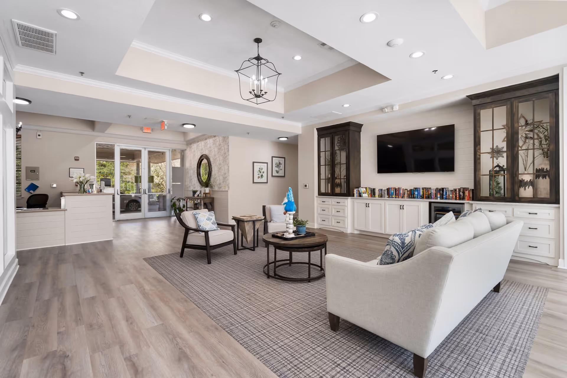 A bright and spacious senior living facility common area with light wood flooring and neutral-colored walls. The room features a white sofa with patterned pillows, two cushioned chairs, and a round coffee table on a large area rug. Built-in cabinets with glass doors flank a wall-mounted flat-screen TV above a fireplace. Books are neatly arranged on the cabinet shelves. In the background, there is a reception desk near glass double doors leading outside, and decorative elements like framed pictures and a round mirror on the walls.