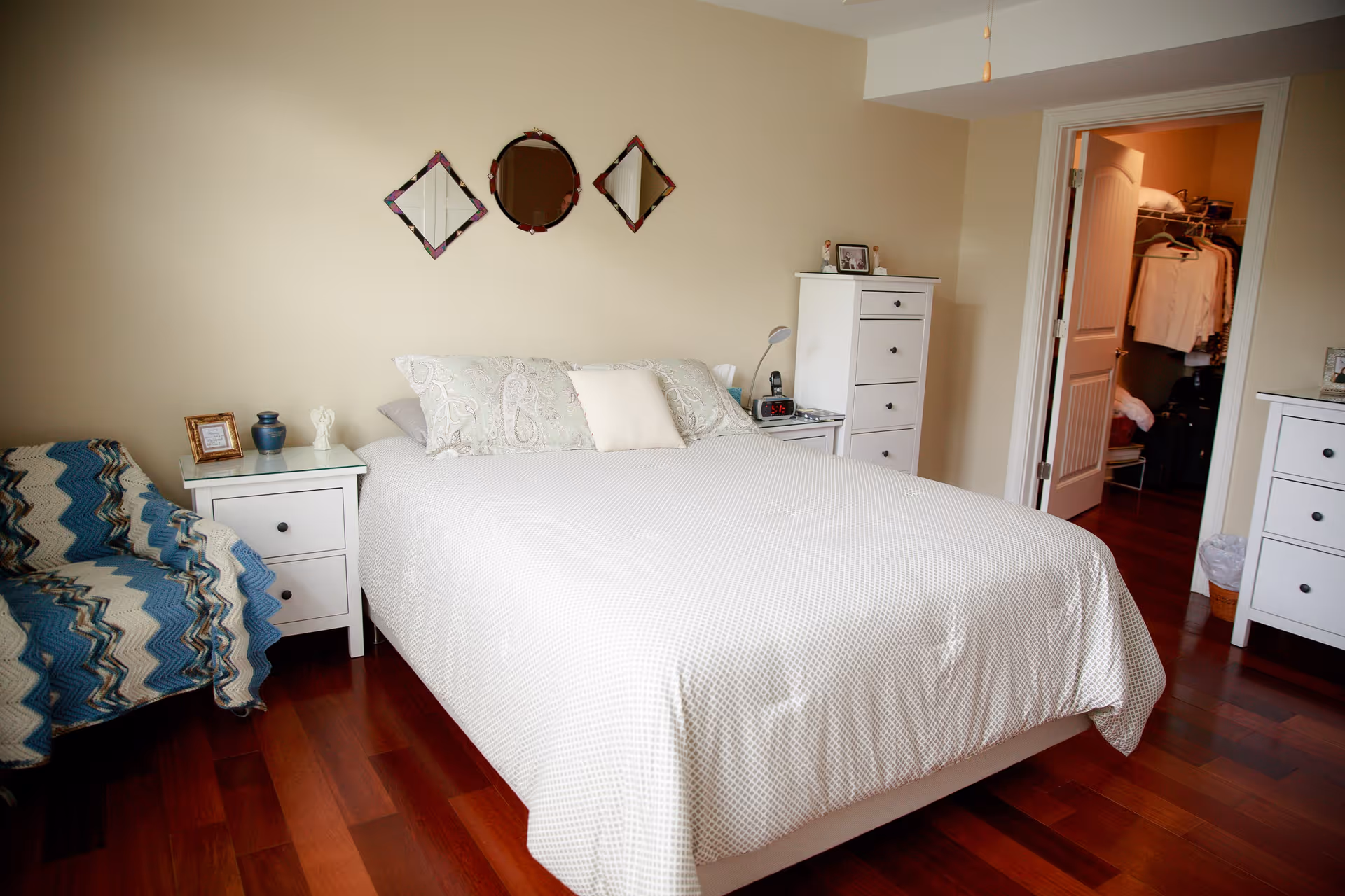 A bedroom with a neatly made bed covered in a white patterned bedspread, two white nightstands on either side of the bed with decorative items, a white chest of drawers, a blue and white patterned armchair, and three decorative mirrors on the wall above the bed. The room has wooden flooring and an open door leading to a walk-in closet.
