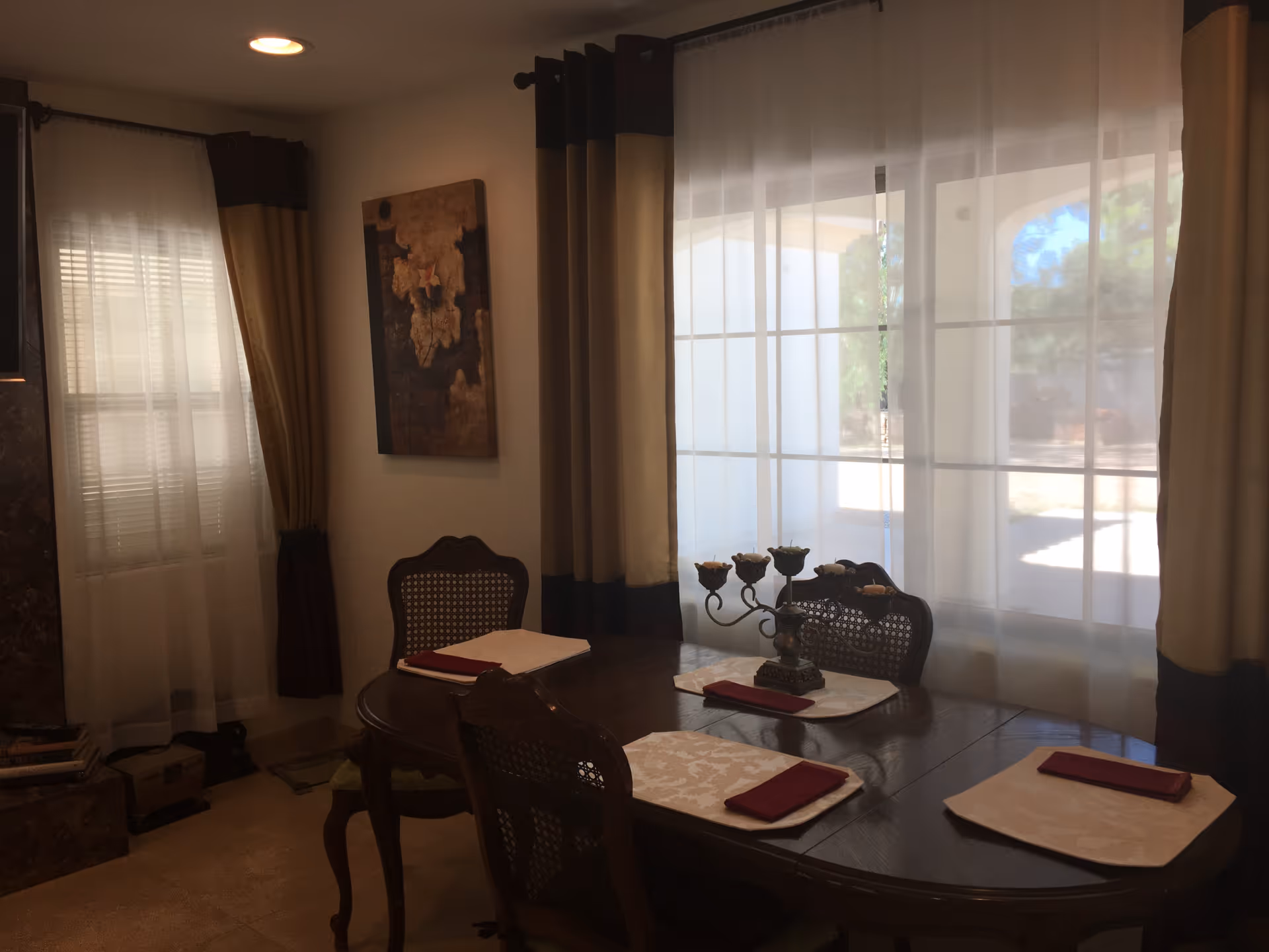 Sunlit dining room with a wooden table set for four, a decorative candelabra centerpiece, and large curtained windows.