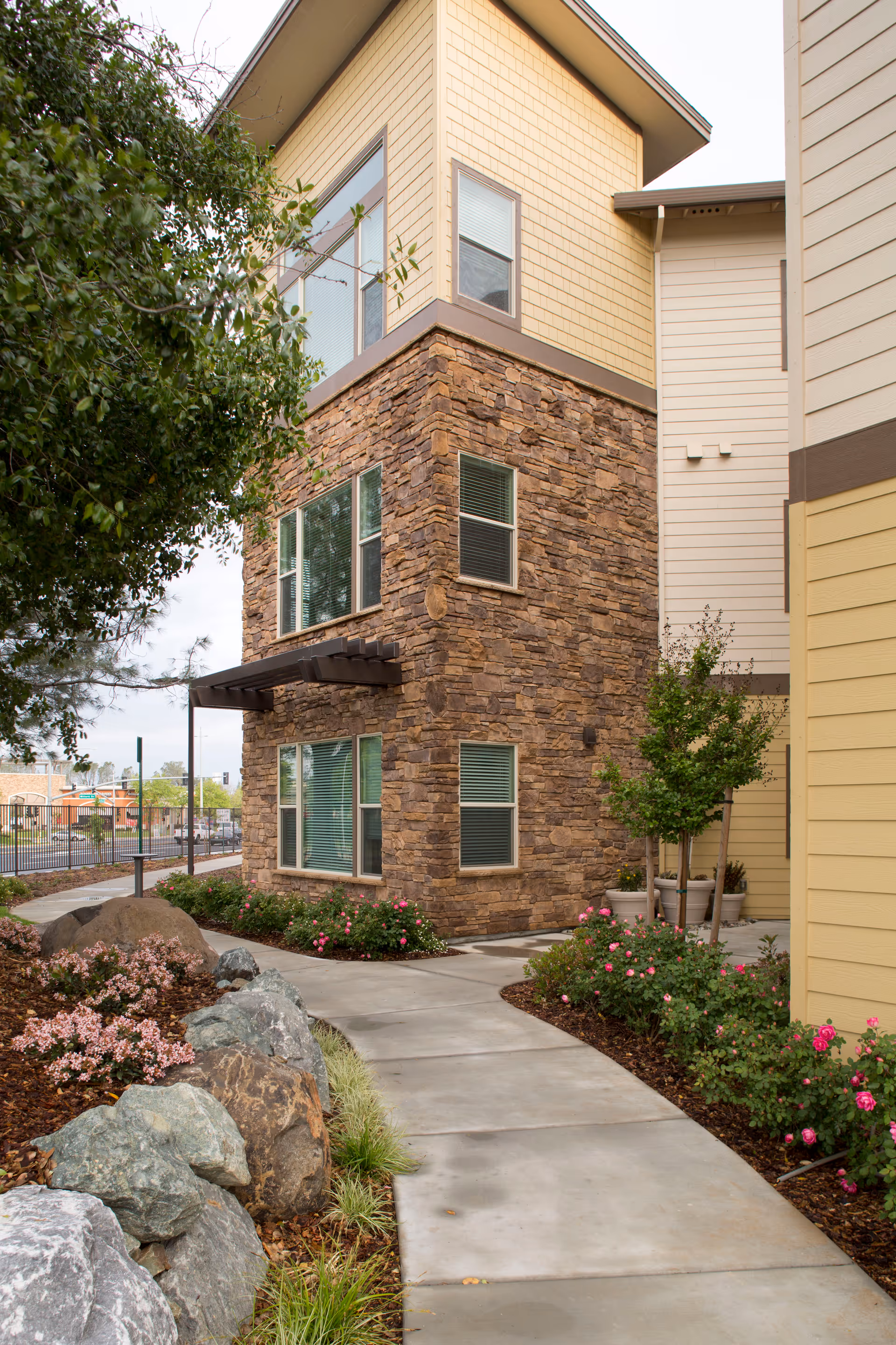 Exterior view of a multi-story building with stone and beige siding, surrounded by landscaped garden beds with rocks, bushes, and flowers along a curved concrete pathway.
