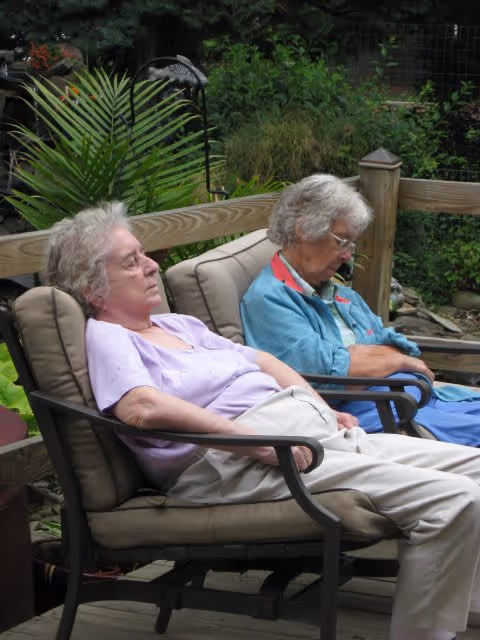 Two elderly women sitting and resting on cushioned outdoor chairs on a wooden deck surrounded by greenery and plants.