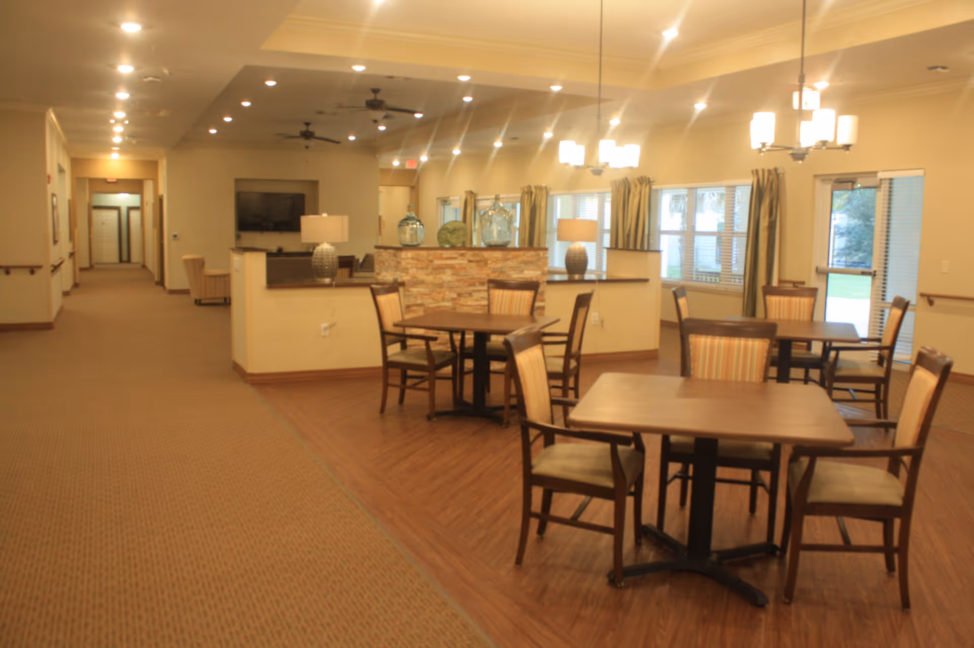 Interior view of a senior living facility dining area with several wooden tables and chairs arranged on a wood floor section. The room has beige walls, ceiling fans, hanging light fixtures, and large windows with curtains letting in natural light. A hallway with carpeted flooring extends to the left, leading to other rooms.