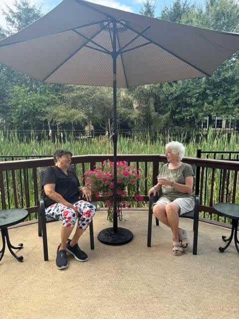 Two elderly women sitting and chatting on chairs under a large patio umbrella on a deck overlooking a pond with tall grasses and trees in the background. There are small round tables on either side of them and a hanging basket of pink flowers in the center.