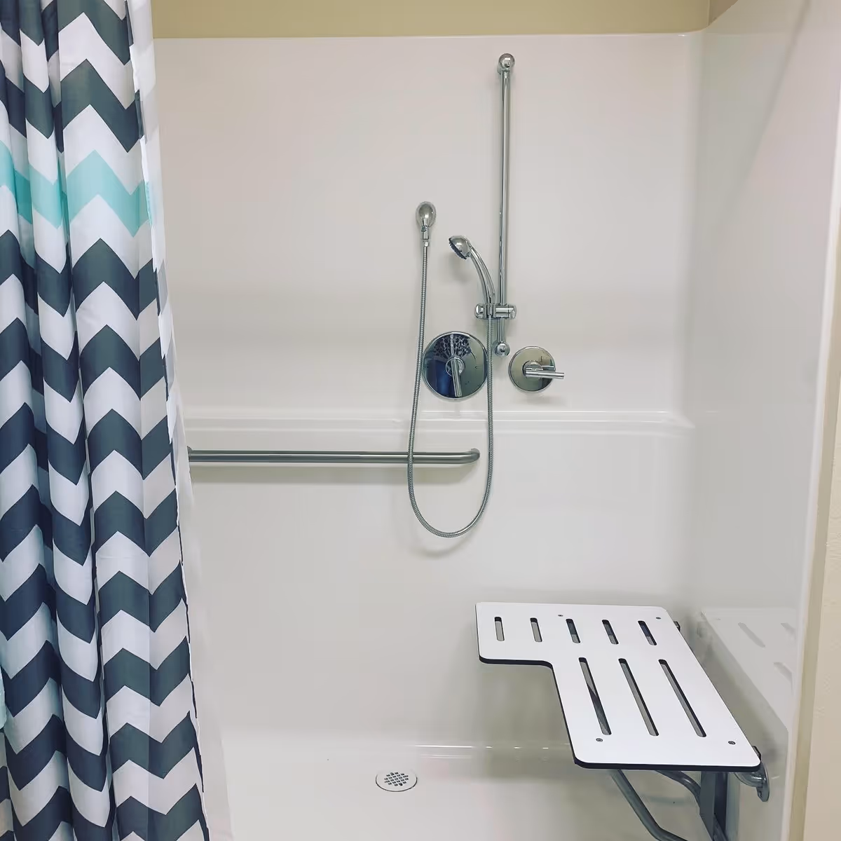 A white accessible shower area with a foldable bench seat, a handheld showerhead attached to a vertical sliding bar, a grab bar on the wall, and a shower curtain with a chevron pattern in shades of black, white, and teal.