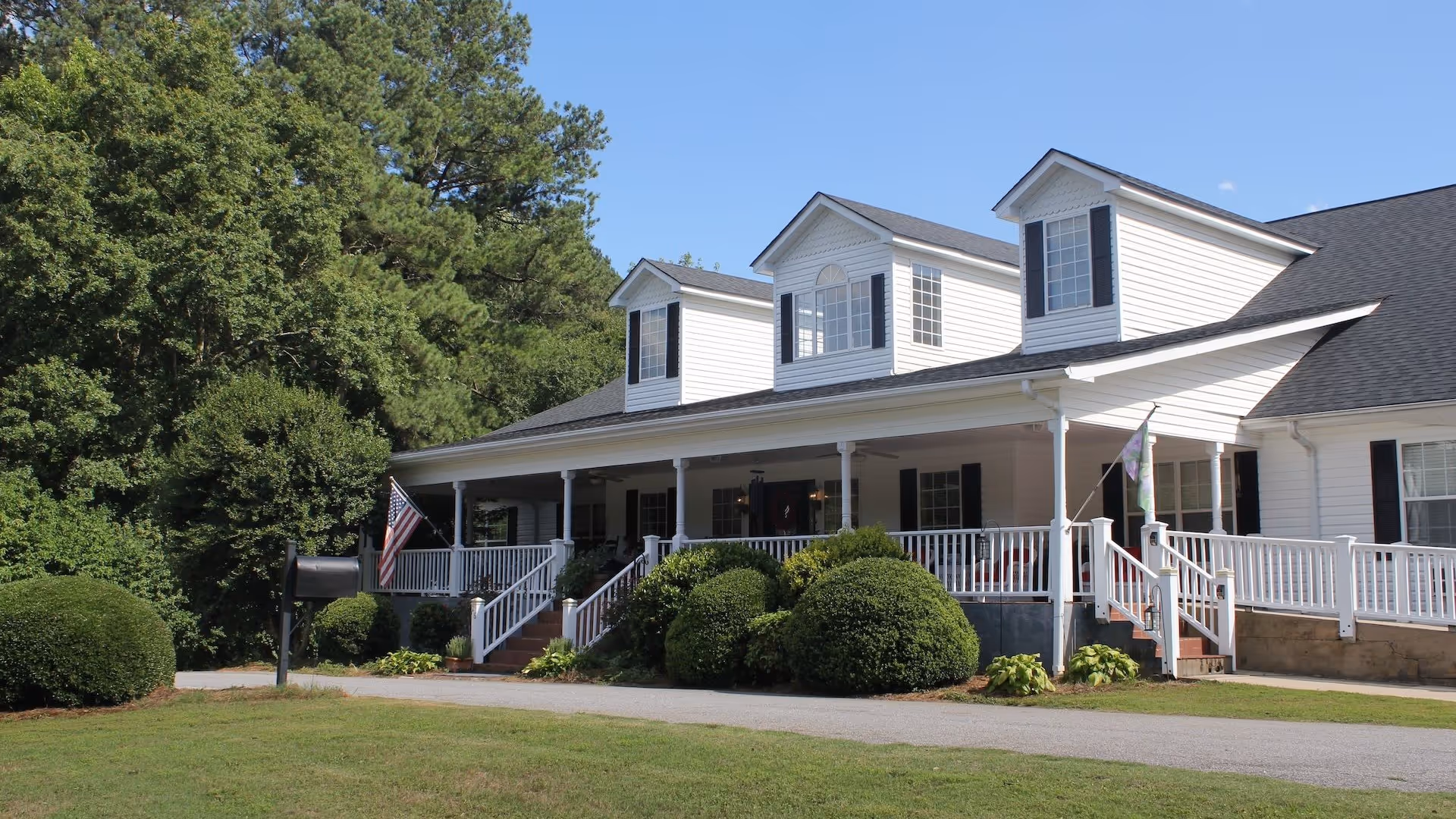 Exterior view of a white two-story building with a large porch, black shutters, and dormer windows. The building is surrounded by green bushes and trees under a clear blue sky. An American flag is displayed near the porch steps.