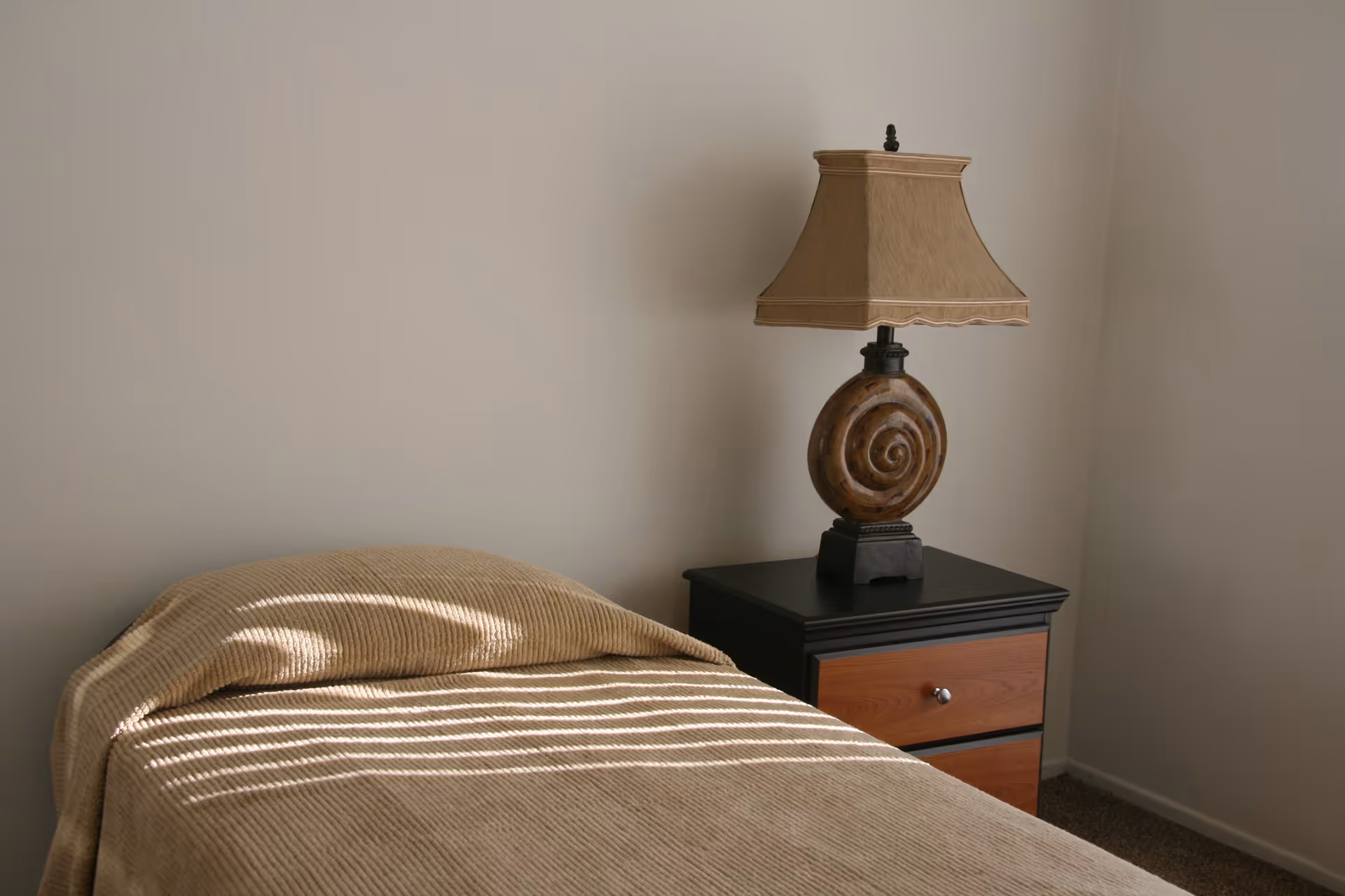A simple bedroom corner with a single bed covered in a beige blanket and a black nightstand with wooden drawers. On the nightstand is a decorative lamp with a beige lampshade and a round, spiral-patterned base. The walls are plain and light-colored.