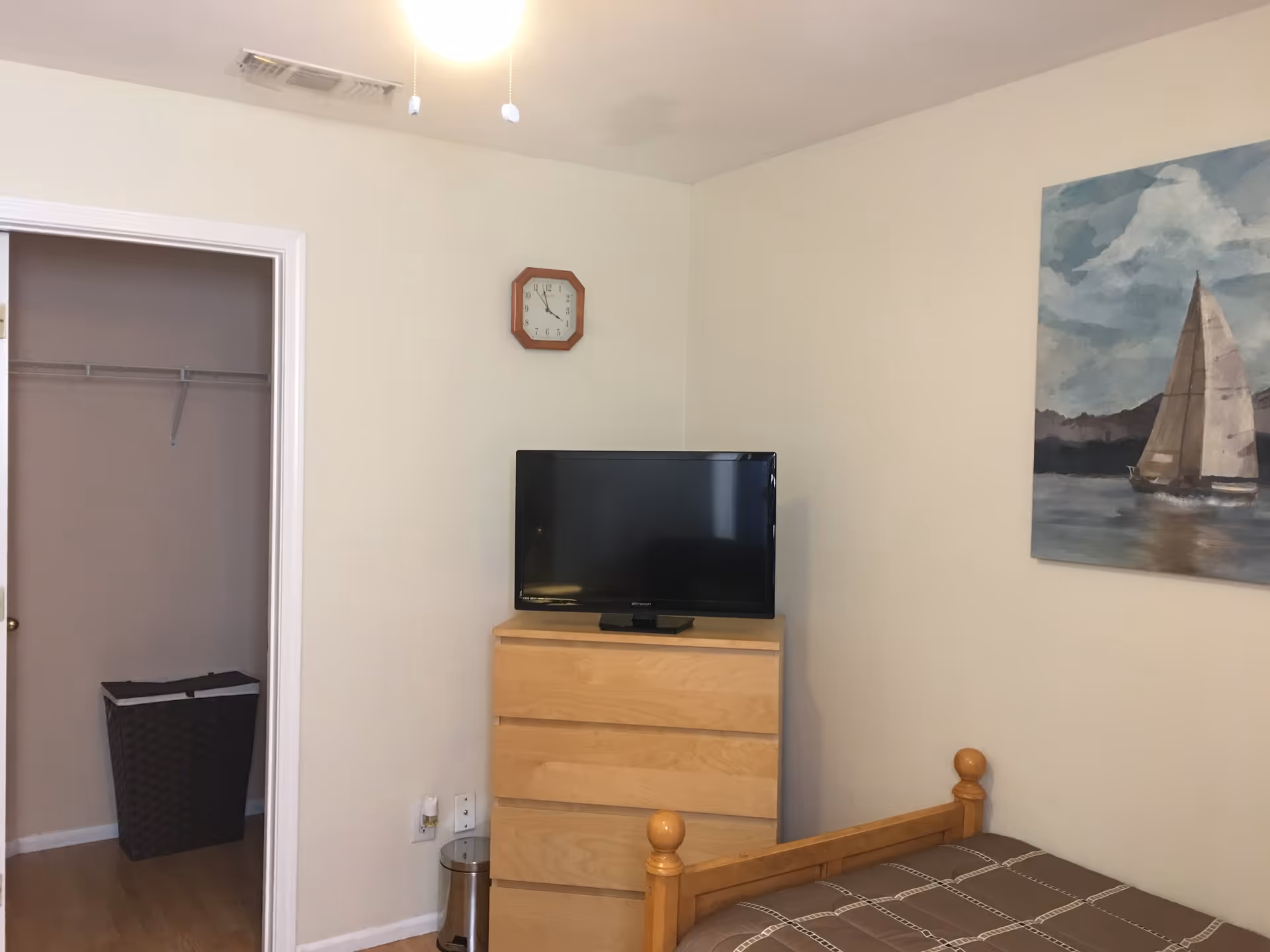 Small bedroom with a wooden bed, dresser topped by a TV, an open closet, and a sailboat painting on the wall.