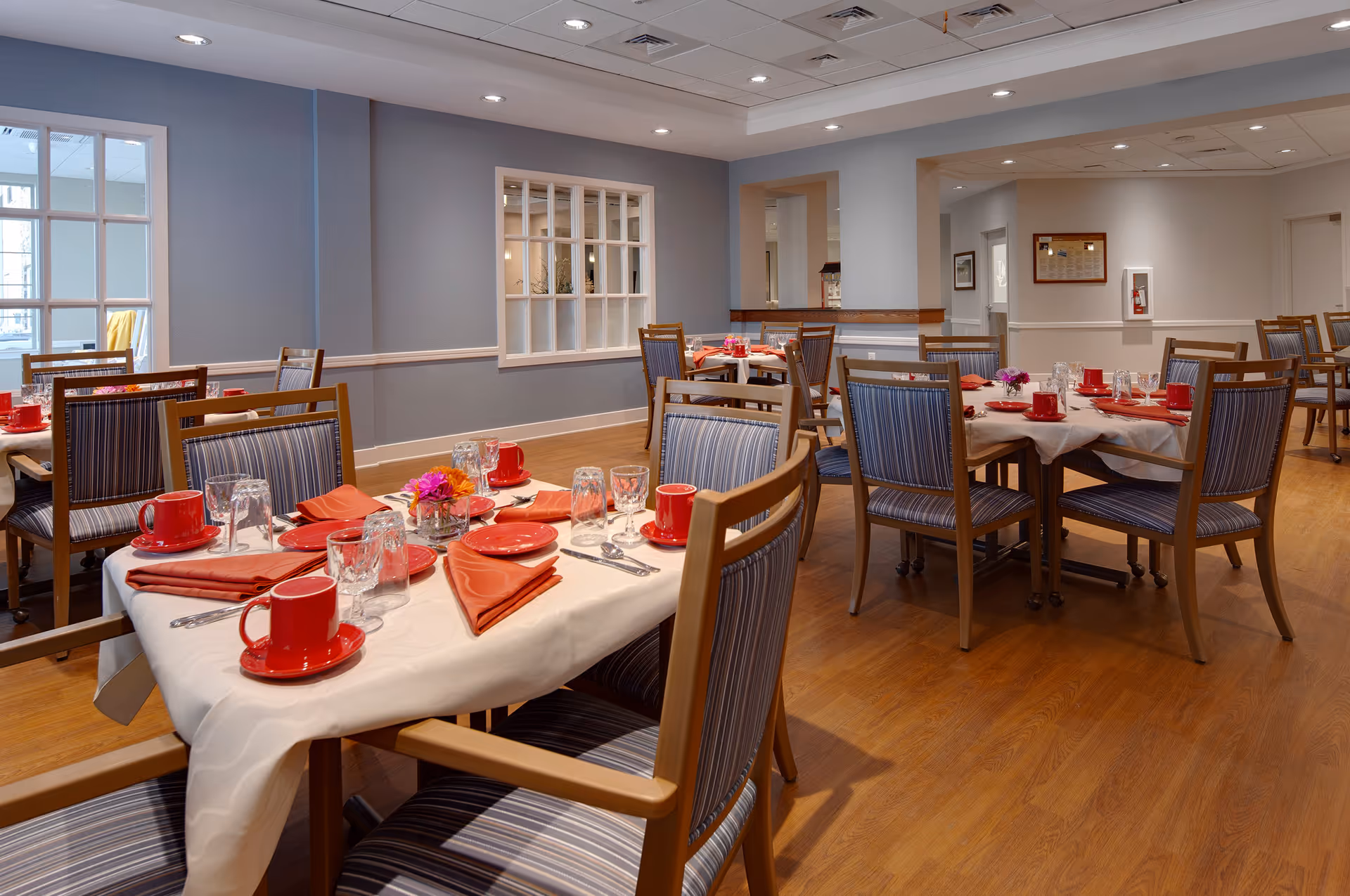 A dining room with several tables set with white tablecloths, red cups and plates, orange napkins, and glassware. The chairs have wooden frames with blue striped cushions. The room has wooden flooring, light blue walls, and recessed ceiling lights.