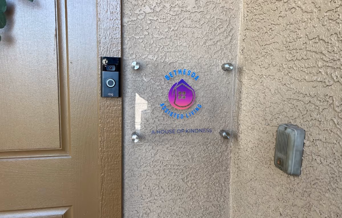 A front door and stucco wall with a clear plaque reading "Bethesda Assisted Living — A House of Kindness," a Ring doorbell, and a vintage doorbell button.