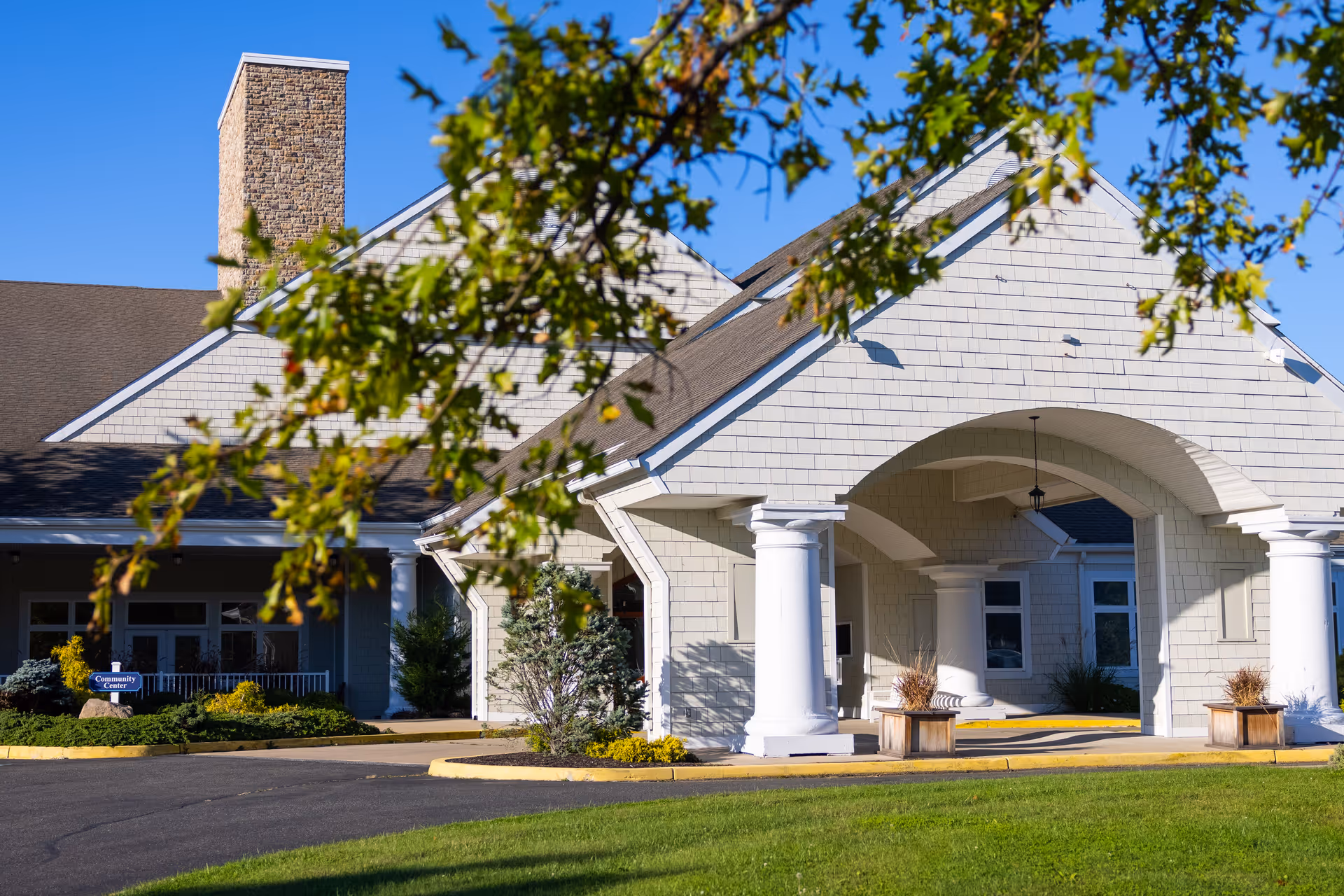 Exterior view of a senior living facility building with a covered entrance supported by large white columns, a stone chimney, and a well-maintained lawn and landscaping under a clear blue sky.